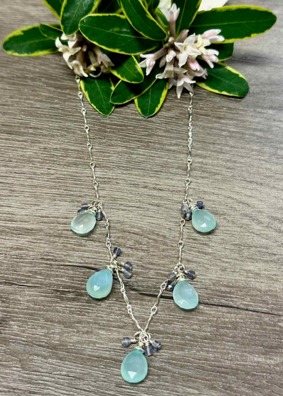 A Chalcedony Drop Necklace with multiple teardrop-shaped stones in light blue color, accompanied by small blue Iolites, strung on a silver chain with a sterling clasp, displayed against a floral background.