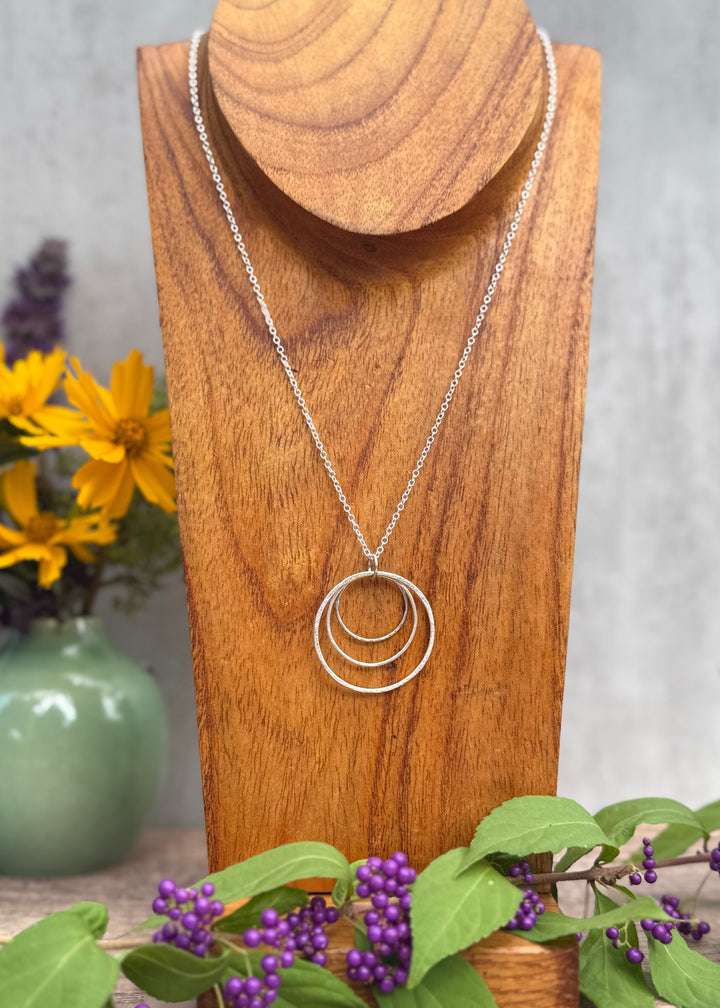 Silver necklace with three interlocking rings on a wooden stand with flowers and greenery in the background