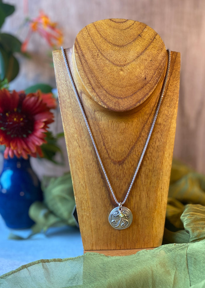 A silver necklace with a circular pendant featuring the Basque cross design, displayed on a wooden stand with a green cloth underneath.